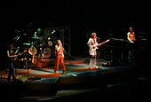 Five men performing on a stage; two are standing behind microphone stands and holding guitars, one is holding a microphone, one is standing behind a keyboard set, and the one furthest back on the stage is sitting behind a drum kit. Lights shine down from above, illuminating a multi-colored stage with geometric shapes.