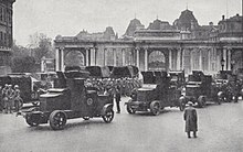 Photographie en noir et blanc d'une file de voitures dans une rue accompagnées d'hommes en tenue militaire.