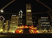 Night view of a bandshell framed by large curved metal surfaces and lit with orange-golden light. A trellis is visible overhead and very tall brightly lit skyscrapers are in the background.