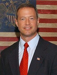 A Caucasian male in a black suit with a light blue shirt and a red tie smiles while standing in front of an American flag.