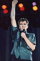 Peter Gabriel mặc áo sơ mi xanh, hát micro trong khi giơ một nắm tay lên trời