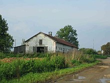 View of a long red-roofed farm building surrounded by overgrown ground at the side of a muddy road