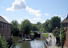 Canal flowing away from the photographer divides into two channels with the right hand one arriving at lock gates. To the is a building and to the right a footpath and a pub.