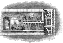 Illustration of a cross section through the Thames Tunnel showing miners on the platforms of a tunnelling shield digging at the face of the excavation whilst others behind on a wheeled platform construct the tunnel lining in brickwork