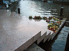 Photo en couleurs (teinte bleutée dominante), prise à Amsterdam en novembre 2003, et montrant le « Homomonument » : un monument dédié aux victimes homosexuelles du nazisme. La photo en montre seulement une partie : un assemblage de cinq dalles en granit rose qui forment un escalier triangulaire permettant d’accéder, du trottoir, au bord d’un canal (de gauche à droite sur la photo). La dernière marche, une plate-forme triangulaire, qui semble flotter sur l’eau du canal, est en partie recouverte de gerbes de fleurs. L’arrière-plan de la photo est constitué par l’étendue d’eau du canal.