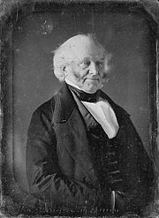 Half-length photographic portrait of an elderly, balding man dressed in a dark coat, vest and cravat