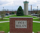 A red brick sign in the foreground says "FCI MILAN". In the back, a solitary green tree is visible atop a grassy mound, with a tower to its right.