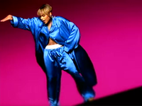 A women showing a white boxer inside her pants, with her right hand up and her left hand on her hip, backed by a dark pink background.