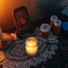 A table with a lit candle, medication pills, a pair of eye glasses, and a small portrait of Mary.