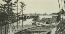 "Black and white photo of a road meandering alongside a river rapids, eventually crossing the top of the rapids along a steel arch bridge."