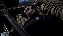 In a darkened room, a deceased man is lying in a coffin. His face is cracked and weathered; the effects of being buried for three months.