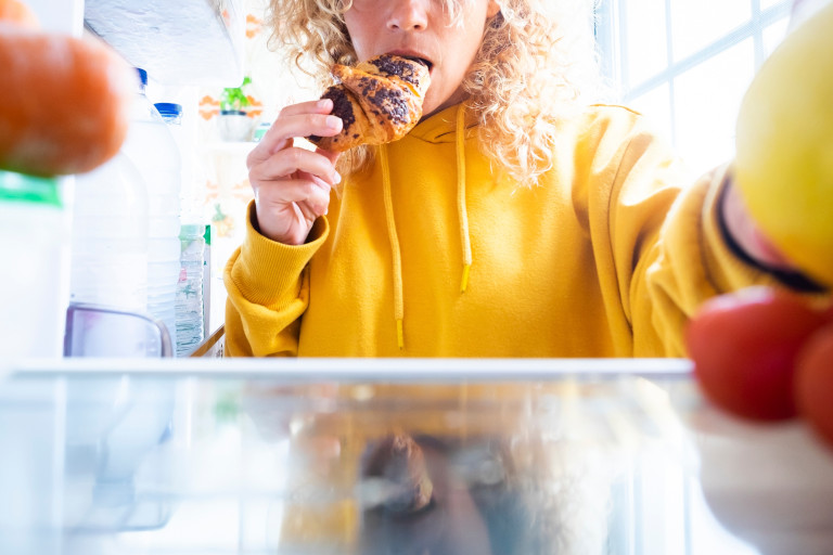 Woman standing in front of the open refrigerator while eating a chocolate croissant and reaching for something else.