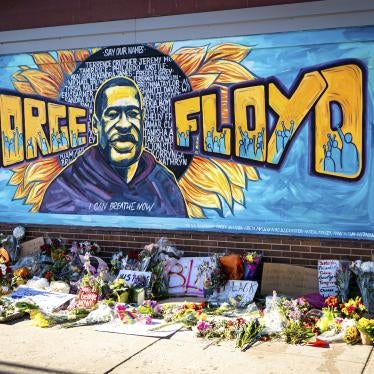 A mural dedicated to George Floyd and honoring the black lives matter movement, showing a depiction of George Floyd's face with his name and supporters of the movement against a blue painted background. Flowers and handwritten signs adorn the sidewalk beneath the mural.