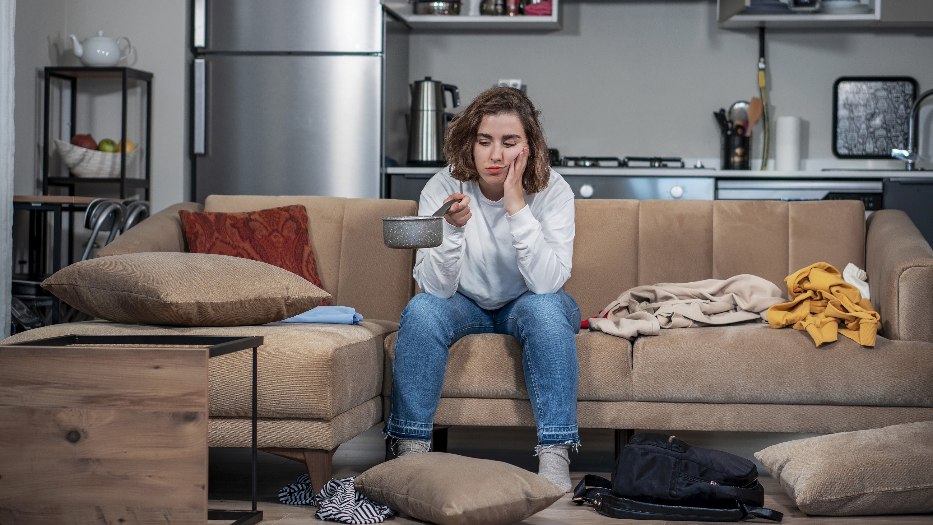 Woman on a sofa in her rental flat holding a saucepan to collect water from the leaking ceiling