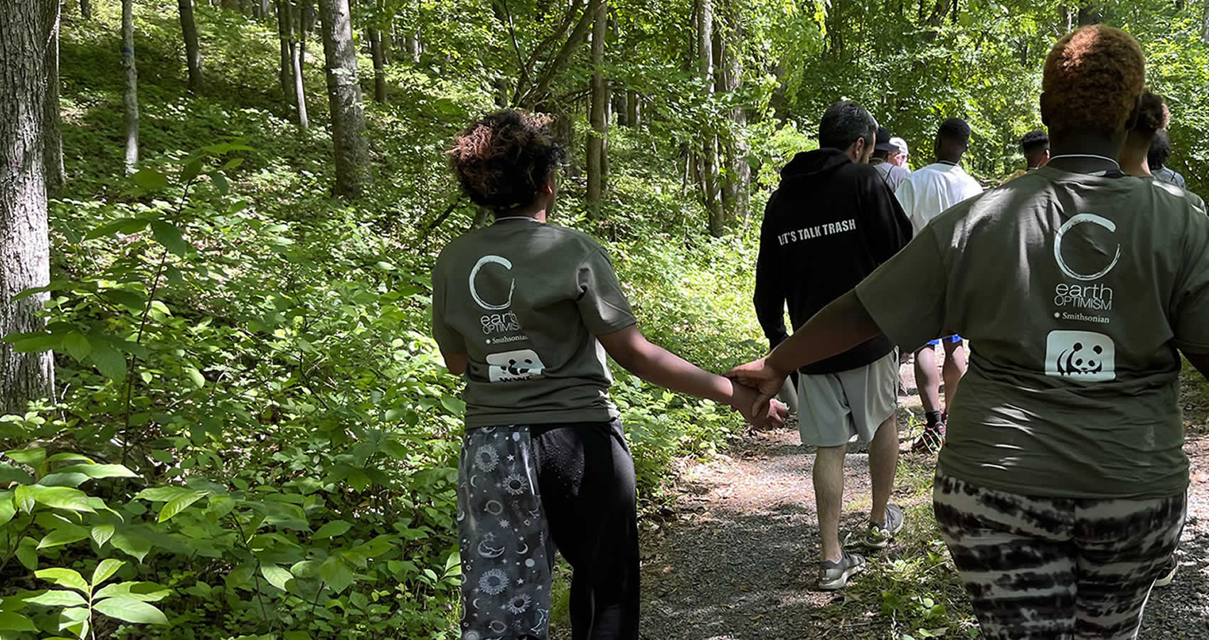 A group of young people walk through a wooded nature path. Two people in the foreground are holding hands. Their t-shirts have white Smithsonian Earth Optimism and World Wildlife Fund logos on them.