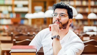 Portrait of young thinking bearded man student with stack of books on the table before bookshelves in the library