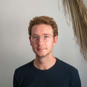 Image of Connor Benedict. Wearing glasses, with a dark shirt, in front of a palm frond.