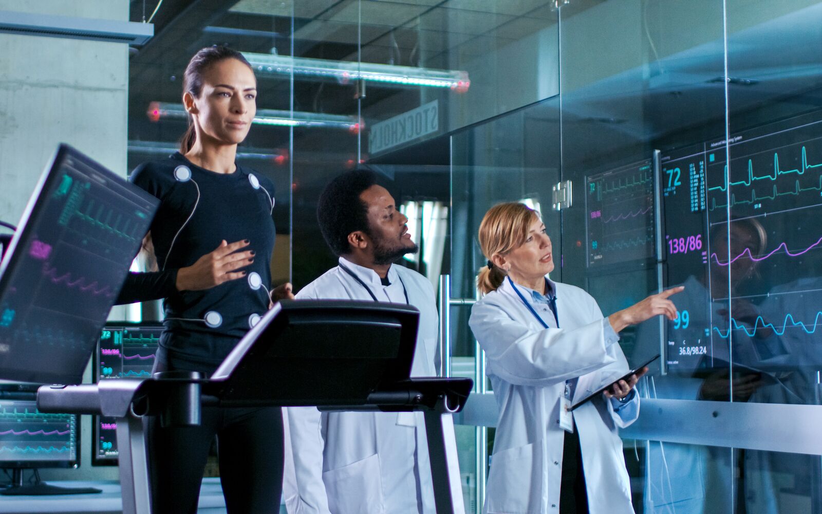 Beautiful Woman Athlete Runs on a Treadmill with Electrodes Attached to Her Body, while Two Scientists Supervise Watching EKG Data Showing on Laboratory Monitors.